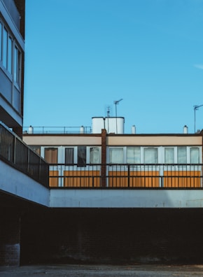 A section of urban architecture featuring a mix of modern and somewhat industrial design elements. The image captures a clear blue sky above a building with alternating orange and white window panels, covered by a balcony or walkway. Metal railings and an exposed brick section add to the linear aesthetic.