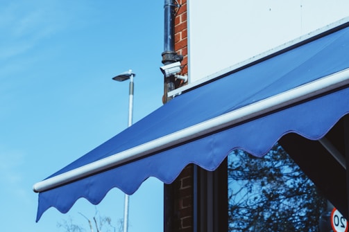 A blue awning is attached to a building with a red brick facade. A security camera is mounted on the corner. The sky is clear and blue, and there is a streetlight in the background.