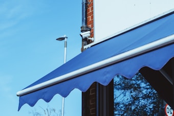A blue awning is attached to a building with a red brick facade. A security camera is mounted on the corner. The sky is clear and blue, and there is a streetlight in the background.