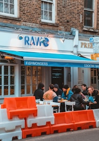 A group of people is seated at an outdoor table of a restaurant with a blue awning labeled 'FISHMONGERS' and 'COCKTAIL BAR'. The restaurant sign reads 'RAW: FISH & COCKTAILS'. Orange and white barriers are placed in front of the restaurant. The building has a brick facade and large windows.