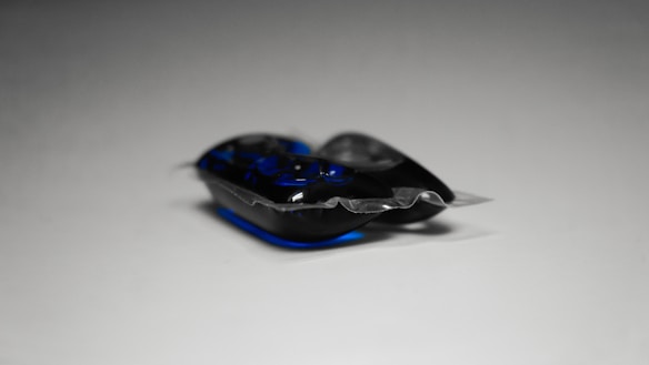A close-up of a single laundry detergent pod with a combination of dark blue and clear gel segments, packaged in a transparent plastic wrapper. The background is a neutral gray, contrasting with the vivid colors of the pod.