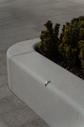 A concrete planter on a tiled pavement contains a small bush with green and yellow leaves. A metal piece is affixed to the edge of the planter.
