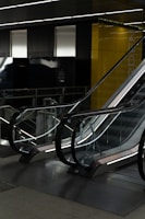 A modern, indoor escalator with reflective glass sides and metallic railings, positioned in a dimly lit environment. Yellow walls and dark panelling surround the area, creating a sleek, contemporary atmosphere.