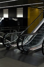 A modern, indoor escalator with reflective glass sides and metallic railings, positioned in a dimly lit environment. Yellow walls and dark panelling surround the area, creating a sleek, contemporary atmosphere.