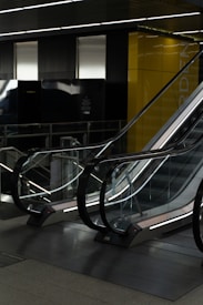 A modern, indoor escalator with reflective glass sides and metallic railings, positioned in a dimly lit environment. Yellow walls and dark panelling surround the area, creating a sleek, contemporary atmosphere.