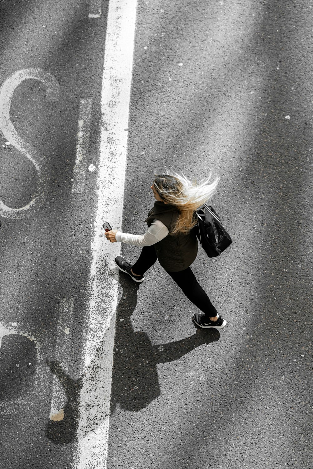 黒いジャケットと黒いズボンを着た女性が、昼間に灰色のアスファルト道路を歩く