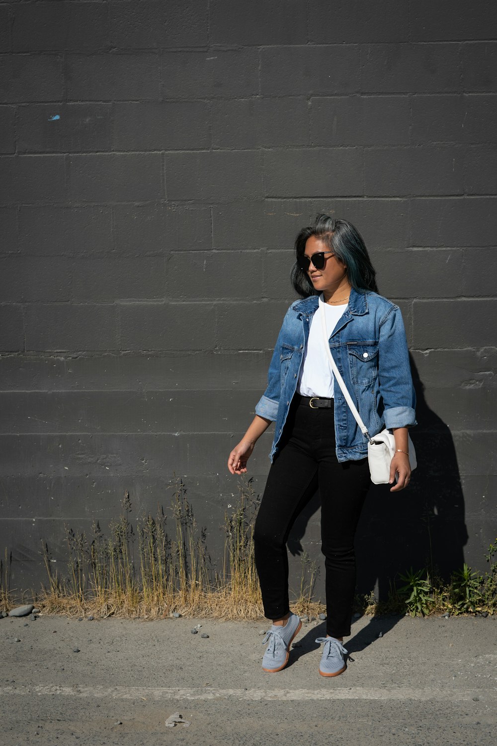 mulher na jaqueta jeans azul e calças pretas de pé ao lado da parede cinza