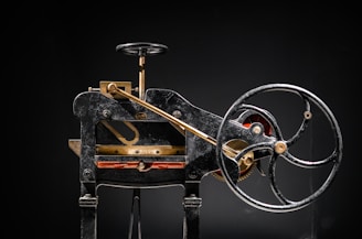A vintage mechanical device composed of mainly black metal with some brass elements. The machine features a large hand wheel, connected via rods and levers, and includes a cutting blade at the base. Intricate components and gears add to the complexity of the machinery.