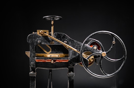 A vintage mechanical device composed of mainly black metal with some brass elements. The machine features a large hand wheel, connected via rods and levers, and includes a cutting blade at the base. Intricate components and gears add to the complexity of the machinery.