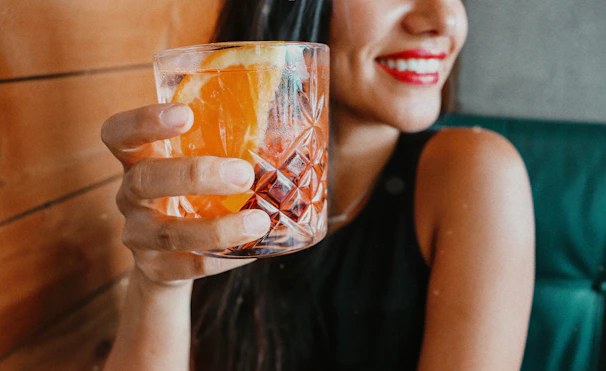 A happy woman holding a glass of bright orange detox juice, smiling with glowing skin in natural sunlight.