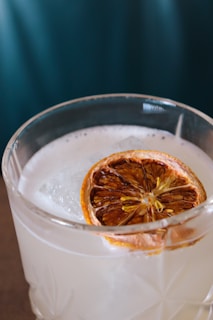 A close-up view of a clear glass filled with a light-colored beverage, topped with a slice of dried citrus fruit and a large ice cube. The drink has a frothy texture on the surface and features an intricate glass design.