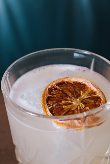 A close-up view of a clear glass filled with a light-colored beverage, topped with a slice of dried citrus fruit and a large ice cube. The drink has a frothy texture on the surface and features an intricate glass design.