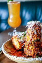A plate featuring grilled corn on the cob coated with a spicy, reddish-orange sauce and sprinkled with shredded cheese. The corn is garnished with herbs, providing a vibrant contrast. In the background, a tall, refreshing drink with an orange hue is visible, set against a blurry, dark teal backdrop.