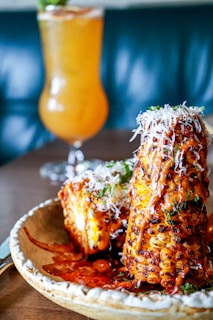 A plate featuring grilled corn on the cob coated with a spicy, reddish-orange sauce and sprinkled with shredded cheese. The corn is garnished with herbs, providing a vibrant contrast. In the background, a tall, refreshing drink with an orange hue is visible, set against a blurry, dark teal backdrop.