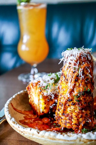 A plate featuring grilled corn on the cob coated with a spicy, reddish-orange sauce and sprinkled with shredded cheese. The corn is garnished with herbs, providing a vibrant contrast. In the background, a tall, refreshing drink with an orange hue is visible, set against a blurry, dark teal backdrop.