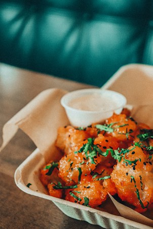 A plate of crispy fried food garnished with fresh green herbs, accompanied by a small container of sauce. The dish is served in a light-colored, biodegradable container and placed on a wooden table. The background has a soft, blurred teal hue.