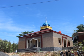 A small, modest mosque with a blue and white dome crowns the building. The structure is mostly rectangular with beige and red-brown walls. The entrance features a simple, arch-shaped design, and there are a few steps leading up to it. Several trees and a clear blue sky are visible in the background, providing a serene atmosphere. A few motorcycles are parked in front of the mosque, indicating its accessibility.