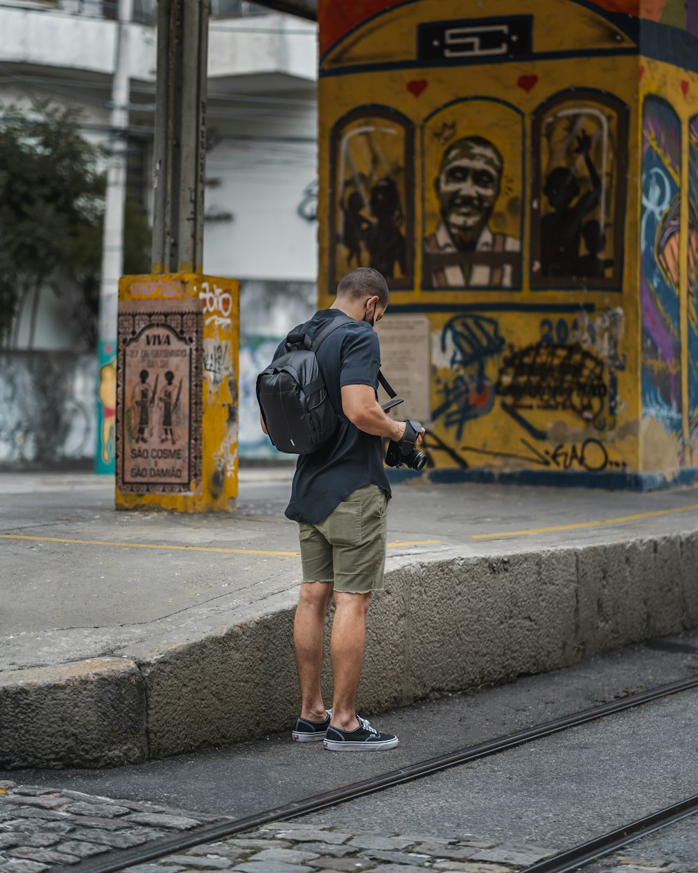 Hombre con camisa negra de manga larga y pantalones cortos marrones de pie en la acera durante el día