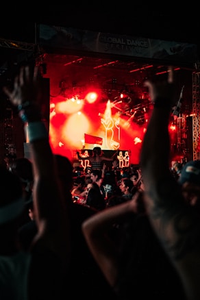 A large crowd of people at a music festival, raising their hands in front of a stage illuminated with bright red and orange lights. Neon outlines of dancers are visible on stage, enhancing the energetic atmosphere. The event is taking place during nighttime.