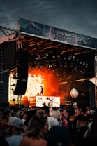 A vibrant music festival scene with a large stage, featuring a performer silhouetted against a backdrop of fiery orange lights. The audience is gathered, enjoying the energetic atmosphere while waving their hands. A large beach ball is visible bouncing among the crowd.