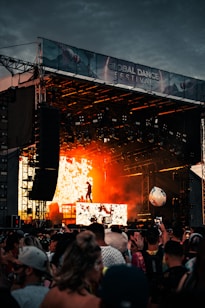 A vibrant music festival scene with a large stage, featuring a performer silhouetted against a backdrop of fiery orange lights. The audience is gathered, enjoying the energetic atmosphere while waving their hands. A large beach ball is visible bouncing among the crowd.