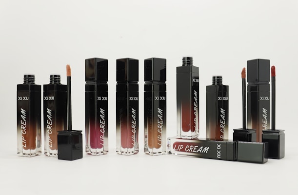 A collection of sleek lip cream containers in various shades, arranged neatly. Some tubes are open showing the applicators.