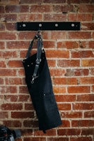 A black apron with metal buckles hangs on a brick wall mounted with a black metal rack featuring round hooks. Below and partially in view is a skateboard with black wheels.