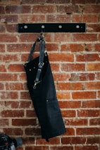 A black apron with metal buckles hangs on a brick wall mounted with a black metal rack featuring round hooks. Below and partially in view is a skateboard with black wheels.