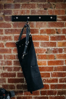 A black apron with metal buckles hangs on a brick wall mounted with a black metal rack featuring round hooks. Below and partially in view is a skateboard with black wheels.
