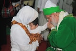 An elder in a green robe and white hat is gently kissing the hand of a baby held by a woman in a white headscarf. They are in a warmly lit environment with blurred individuals in the background.