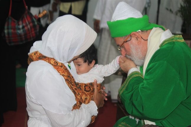 An elder in a green robe and white hat is gently kissing the hand of a baby held by a woman in a white headscarf. They are in a warmly lit environment with blurred individuals in the background.