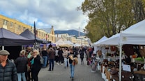 People are gathered at an outdoor market with numerous stalls lined up on either side of a pedestrian path. Tents cover the stalls, which are displaying various goods such as bags and handcrafted items. The scene is set amidst historic buildings and lush trees, with a mountain visible in the background under a partly cloudy sky.