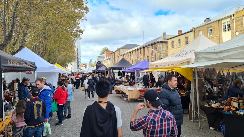 A bustling outdoor market with various stalls set up under white and colorful tents. People are walking along the cobblestone path, browsing and interacting with vendors. Trees with autumn foliage line one side of the street, while historic-style buildings provide a backdrop on the other.