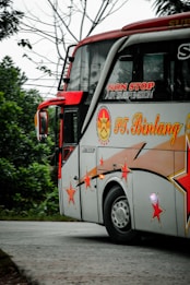 A decorated bus with vibrant red and orange star designs is traveling on a road surrounded by greenery. The bus has branding with text indicating non-stop air suspension. It is mainly white with red accents and features large windows.