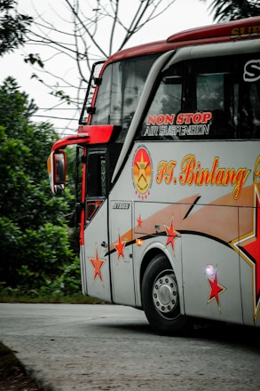 A decorated bus with vibrant red and orange star designs is traveling on a road surrounded by greenery. The bus has branding with text indicating non-stop air suspension. It is mainly white with red accents and features large windows.