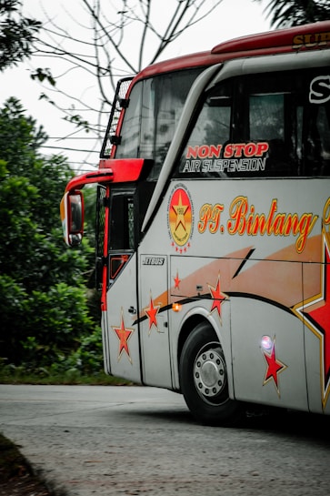 A decorated bus with vibrant red and orange star designs is traveling on a road surrounded by greenery. The bus has branding with text indicating non-stop air suspension. It is mainly white with red accents and features large windows.