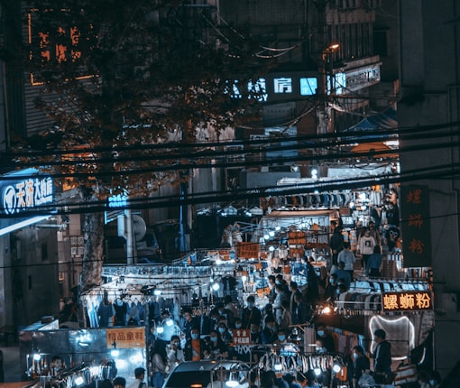 A bustling night market is filled with people and various stalls, illuminated by bright neon signs in Chinese characters. The scene is crowded, with a diverse range of goods displayed under the glow of small overhead lights.