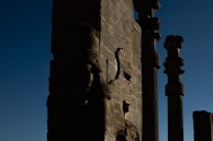 The image features ancient stone carvings and tall columns against a deep blue sky. The carvings appear to depict mythical creatures or figures, showcasing intricate craftsmanship typical of ancient architecture.