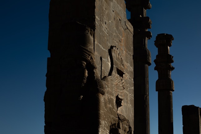 The image features ancient stone carvings and tall columns against a deep blue sky. The carvings appear to depict mythical creatures or figures, showcasing intricate craftsmanship typical of ancient architecture.