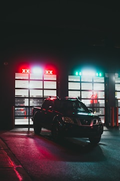 A black pickup truck is parked in front of an illuminated garage entrance at night. The garage has two sections with bright red and green lights above, casting reflections on the wet ground. The setting suggests an automotive facility or car wash.