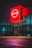 A brightly lit Nissan dealership with a prominent red sign displaying the brand name. The large glass windows reflect ambient light, and the pavement in front shows purplish hue from the reflections. The interior appears empty with visible parking spaces outside.