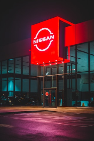 A brightly lit Nissan dealership with a prominent red sign displaying the brand name. The large glass windows reflect ambient light, and the pavement in front shows purplish hue from the reflections. The interior appears empty with visible parking spaces outside.