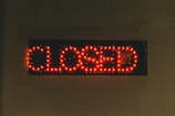 A rectangular black sign displays the word 'CLOSED' in bright red LED lights. The lights stand out against the dark background, creating a clear and attention-grabbing message. The dots formed by the LED lights give the text a digital appearance.