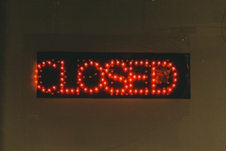 A rectangular black sign displays the word 'CLOSED' in bright red LED lights. The lights stand out against the dark background, creating a clear and attention-grabbing message. The dots formed by the LED lights give the text a digital appearance.