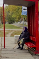 An elderly person is sitting at a red bus stop shelter with a walking stick resting on their lap. The background features a park-like setting with green and yellow trees, suggesting an autumn environment. A blue bus stop sign with schedule details is visible above the person, and a trash can is placed nearby. The person appears to be waiting patiently, dressed warmly in a coat and a purple headscarf.