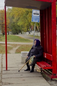 An elderly person is sitting at a red bus stop shelter with a walking stick resting on their lap. The background features a park-like setting with green and yellow trees, suggesting an autumn environment. A blue bus stop sign with schedule details is visible above the person, and a trash can is placed nearby. The person appears to be waiting patiently, dressed warmly in a coat and a purple headscarf.
