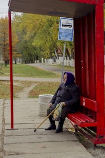 An elderly person is sitting at a red bus stop shelter with a walking stick resting on their lap. The background features a park-like setting with green and yellow trees, suggesting an autumn environment. A blue bus stop sign with schedule details is visible above the person, and a trash can is placed nearby. The person appears to be waiting patiently, dressed warmly in a coat and a purple headscarf.