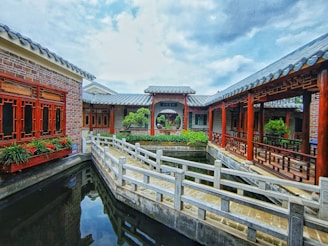 A traditional Chinese architectural setting featuring wooden structures with intricate red woodwork and tiled roofs. A stone bridge crosses a calm body of water, leading to a courtyard with lush vegetation. The sky above is partly cloudy, adding depth to the setting.