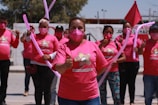 A group of people wearing pink shirts and masks participate in a demonstration or event. They are holding pink balloons and giving thumbs up gestures. A red flag is visible in the background along with trees and part of a building. The shirts have a logo or graphic on them.