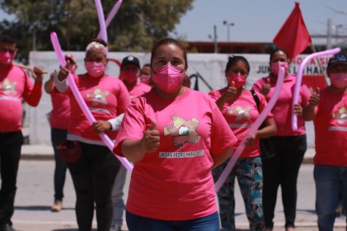 A group of people wearing pink shirts and masks participate in a demonstration or event. They are holding pink balloons and giving thumbs up gestures. A red flag is visible in the background along with trees and part of a building. The shirts have a logo or graphic on them.